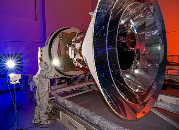NASA's SPHEREx observatory inside a cleanroom oriented in a horizontal position, revealing all three layers of photon shields as well as the telescope. This photo was taken at BAE Systems in Boulder, Colorado, in April 2024. A worker stands next to the Spherex observatory while shining a handheld light towards it