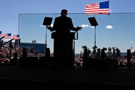 Republican presidential nominee and former president Donald Trump, photographed from behind in silhouette, speaks from behind a glass barrier. In the distance beyond the silhouette of Trump, podium and stage, the audience and several American flags can be seen